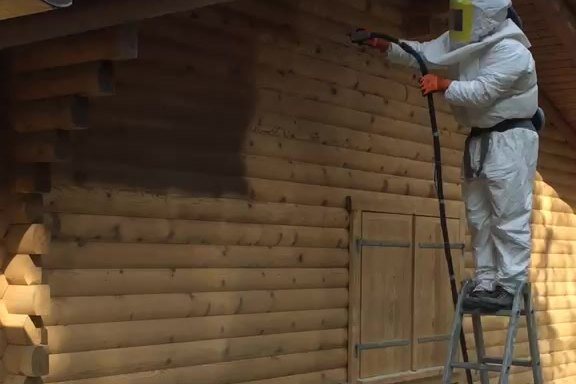 Opérateur en combinaison de protection passant la buse d'aérogommage sur le bardage en rondins d'un chalet